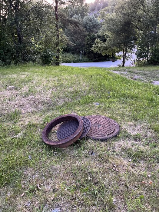 Två brunnsringar med lock ligger på en gräsplan intill en väg omgiven av träd.