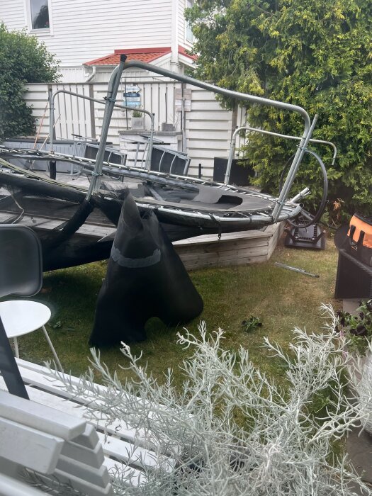 Stormskadad studsmatta med ram och nät på en gräsmatta, omgiven av trädgårdsmöbler och växter, framför vit träbyggnad.