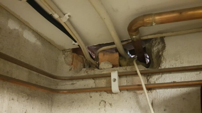 Hål i källarvägg med synliga rör och elkablar, omgiven av murbruk och tegel.