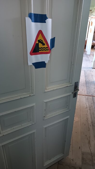 En vit dörr med en varningsskylt för bil vid brygga fasttejpad. Bakom dörren syns ett golv av träplankor.
