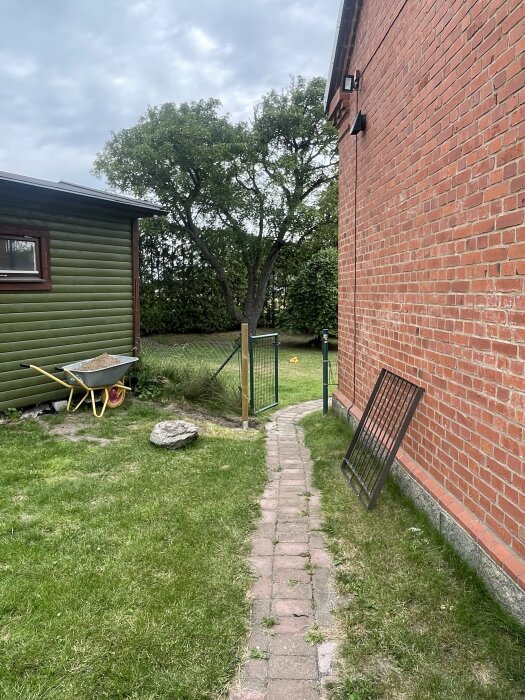 Gångväg mellan röd tegelvägg och grön byggnad med ny grindstolpe, skottkärra bredvid och träd i bakgrunden.