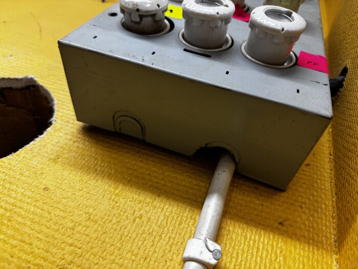 En grå el-central med tre keramiska säkringar, placerad på ett gult underlag med ett hål intill och en vit kabel ansluten undertill.