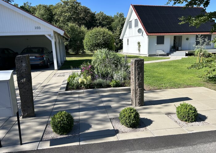 Granitstolpar står på en stenlagd uteplats omgivet av planterade buskar och blommor, med ett vitt hus och grönska i bakgrunden.