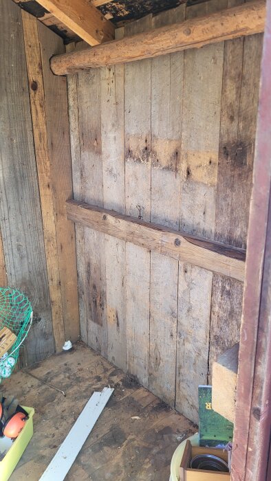 Interiör av ett gammalt utedass med slitna träväggar, en träbalk, och diverse föremål i hörnet.