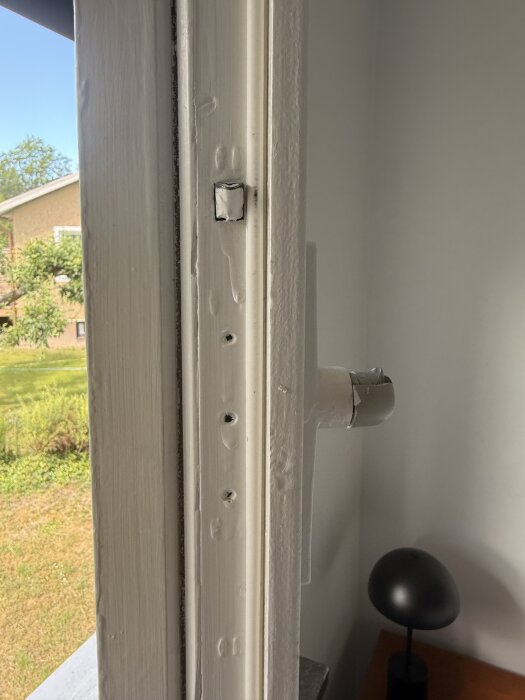 Fönsterbåge med borttagen färg och hål, inomhusmiljö med synlig trädgård i bakgrunden.