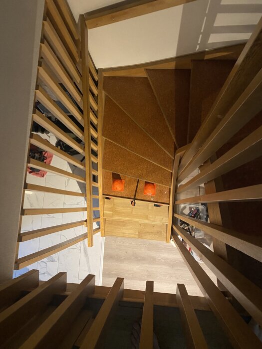 Vy ovanifrån av en spiraltrappa med trädetaljer. Trappstegen har mörk beläggning, omgivna av ljusare ekytor och räcken.