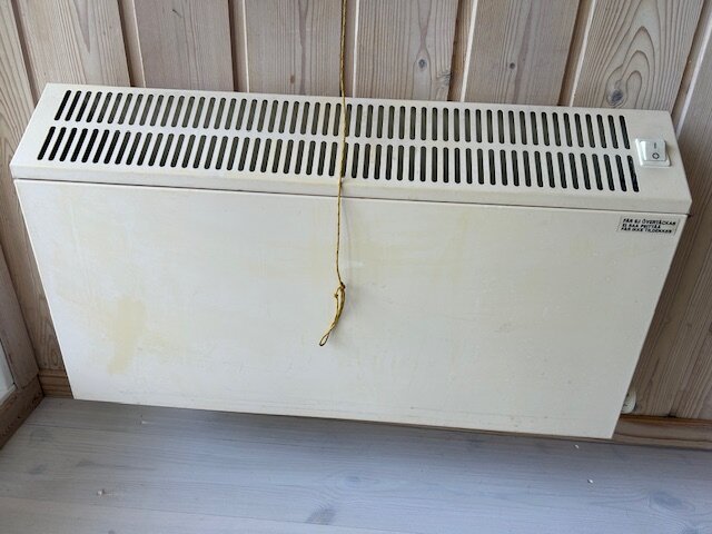 Direktverkande fläktkonvektorelement monterat på trävägg i sommarstuga utan synlig stickpropp, med gul sladd och strömbrytare upptill.