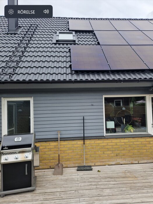 Solpaneler installerade på taket av ett hus med grå fasad och en grill på trädäcket framför.