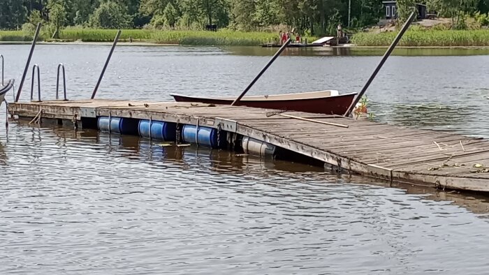 Flytbrygga med blå tunnor under, skev och lutande, med båt och brädor ovanpå.