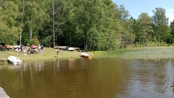 En grupp personer samlade vid en sjö, med grönska och båtar i bakgrunden, en ensam roddbåt synlig vid strandkanten.