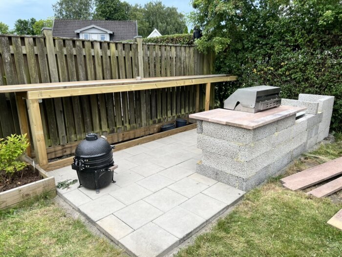 Utomhuskök under uppbyggnad med stengolv, träbänk och grill; betongblock formar grillens struktur, omgiven av plank och grönska.