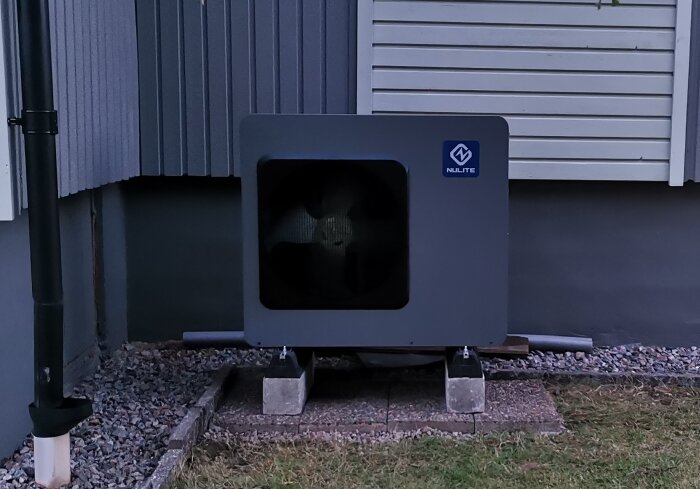 Värmepump installerad utomhus på ett betongfundament vid väggen av ett hus, med synliga rör och omgivande gräsyta.