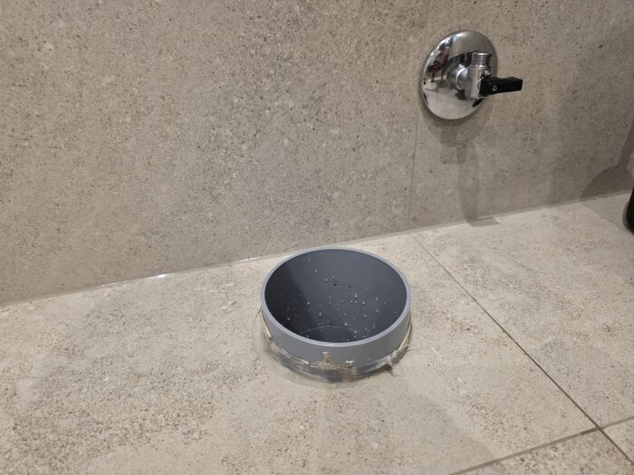 Grå packning kring toalettavloppsrör på beige kakelgolv med synlig vattenventil på väggen.