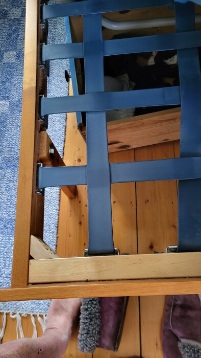 En sängbotten med blå resårband monterade i rutmönster på en träram, synligt golv och en person i tofflor bredvid.