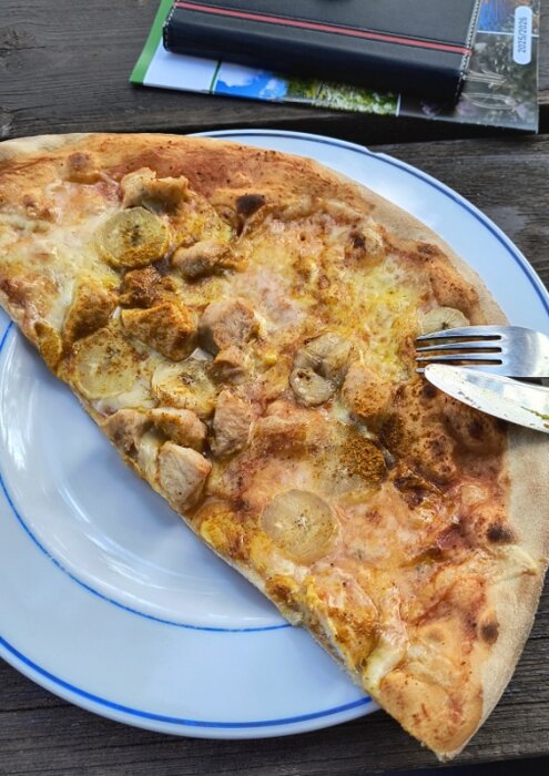 Pizza slice on a plate with banana, chicken, and curry spices, placed on a wooden table alongside a notepad and papers.