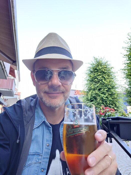 Man i hatt och solglasögon skålar med ölglas utomhus, redo för festival.