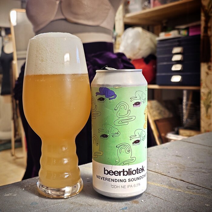 Krus med skummande öl bredvid burk med etiketten "beerbliotek Neverending Soundcheck DDH NE IPA 6.5%".