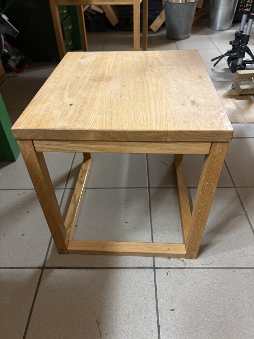 Ekfärgat sidobord i modern, enkel design, med fyrkantiga ben och tvärgående ramar, står på golvplattor i ett verkstadsutrymme.