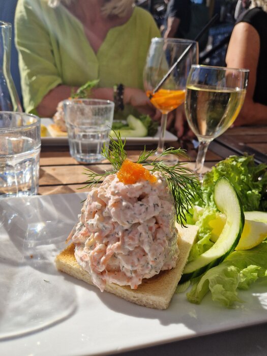 Skagen-torn med dill och löjrom på rostat bröd, serverat med sallad och gurka, i solljus på restaurang i Stockholm.