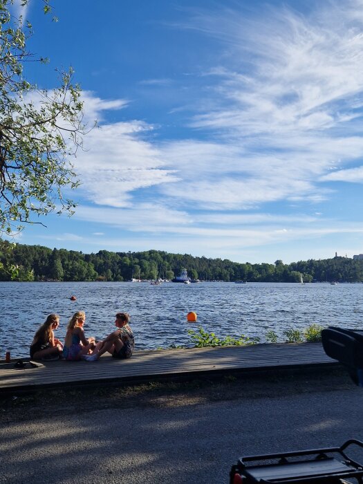 Tre personer sitter vid vattnet i Tanto, Stockholm, under en blå himmel med vita moln.