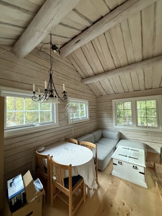 En ljus inredning i ett gästhus med träpanel, en rund matplats, soffhörna och låda under fönster med utsikt över träd.