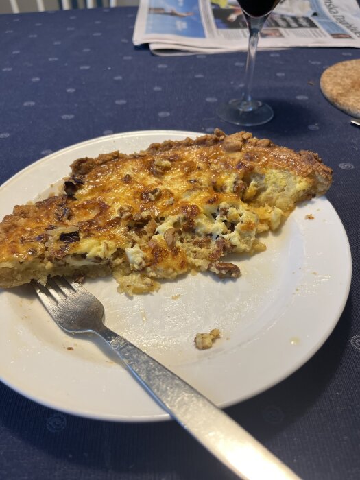 Tallrik med halväten paj, gaffel och kniv på en duk. Bakom syns ett glas rött vin och en tidning.