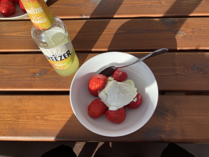 Jordgubbar och glass i en skål på ett träbord bredvid en flaska mango-dryck.