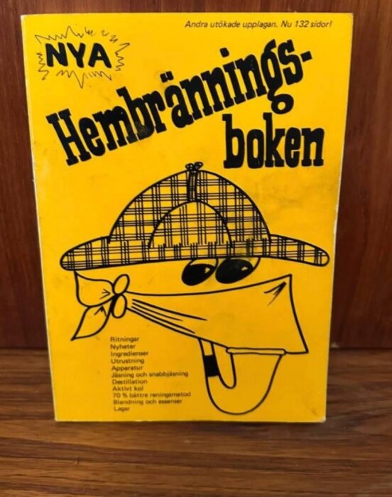 Gul bok med texten "Nya Hembränningsboken", illustrerad med ansikte i hatt och bandana. Listar innehåll som ritningar och ingredienser.