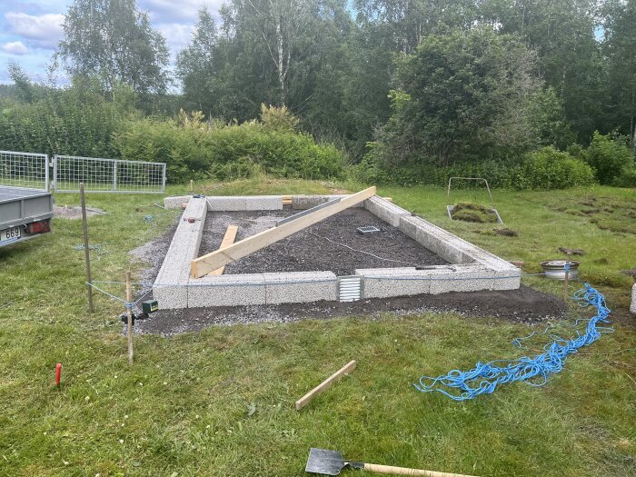 Grundläggning av ett byggprojekt på gräsmatta med murblock, grusfyllning och träbjälkar, omgivna av träd och buskar.