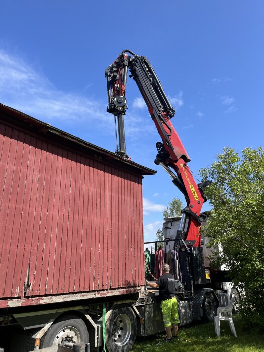 Röd kranbil flyttar en röd byggnad på en solig dag, arbetare i gula byxor övervakar processen.