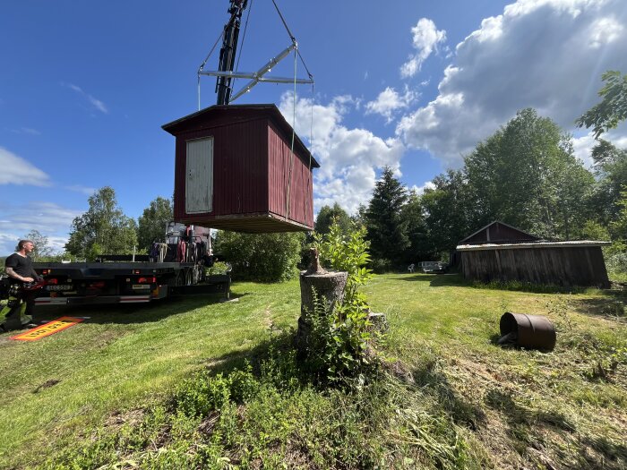 Lyft av en röd stuga med vit dörr från en lastbil i en grönskande trädgård med träd och en lada i bakgrunden.