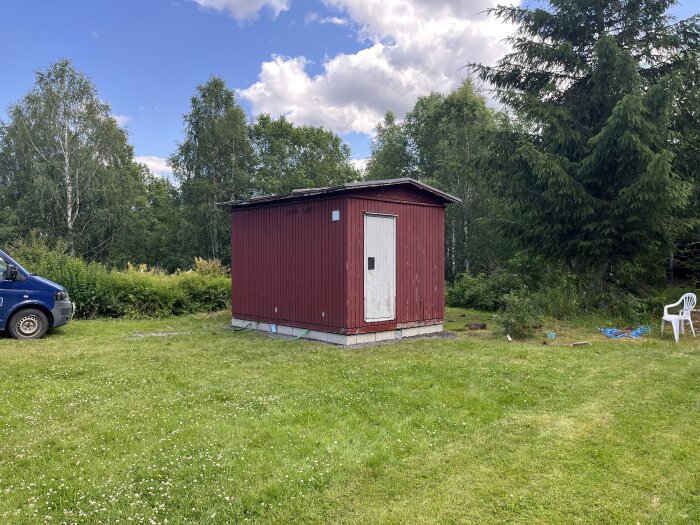 Rött skjul med vit dörr på gräsplan, omgiven av träd och en vit plaststol.