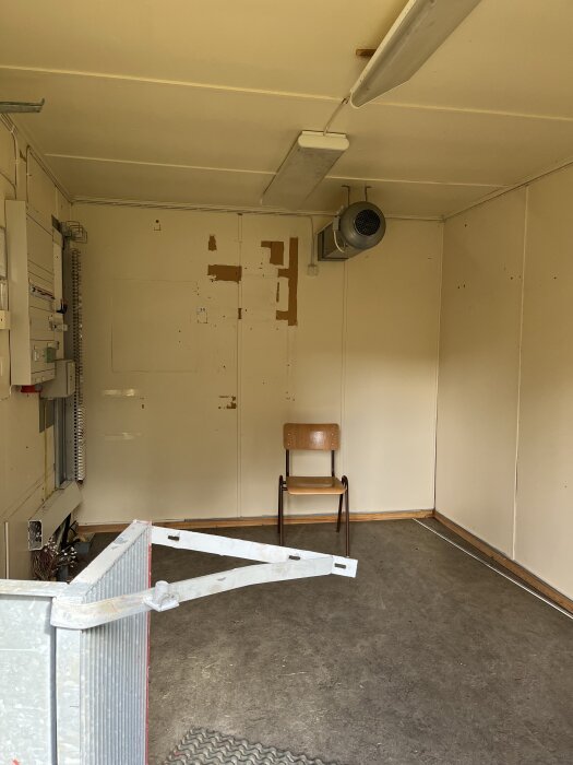 Tomt rum med beige väggar, en ventilationsfläkt, en stol och ett proppskåp på vänstra väggen. Resultatet av ett renoveringsprojekt.