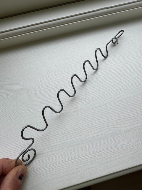 Metallkrok med vågig design placerad på en fönsterbräda. Kroken liknar en stormhasp och har en tydligt snurrad ände.