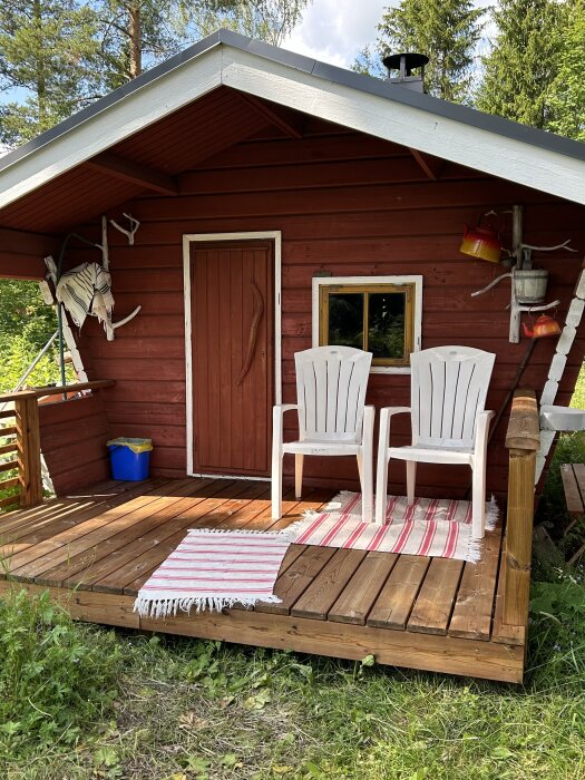Röd bastu med vita plaststolar och trasmattor på verandan, omgiven av grönska.