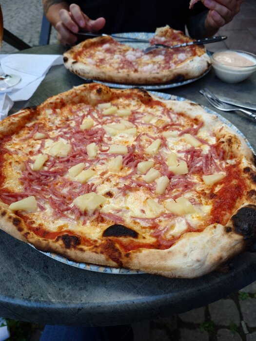 Napolitansk Hawaii-pizza toppad med skinka och ananas på ett bord, med en annan pizza och bestick i bakgrunden.