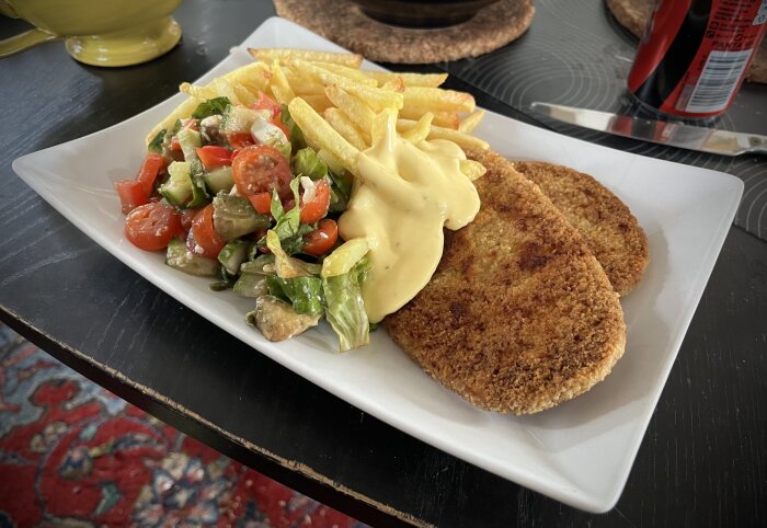 Tallrik med schnitzel, pommes frites, sallad och sås på ett bord.