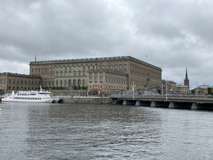 Stort stenbyggnad vid vatten med en vit båt ankrad framför, och en bro som sträcker sig över vattnet under en mulen himmel.