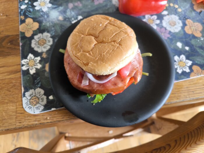 Hamburgare med stor köttbit och smått bröd, toppad med sallad, paprika, rödlök och dressing, serverad på en svart tallrik.
