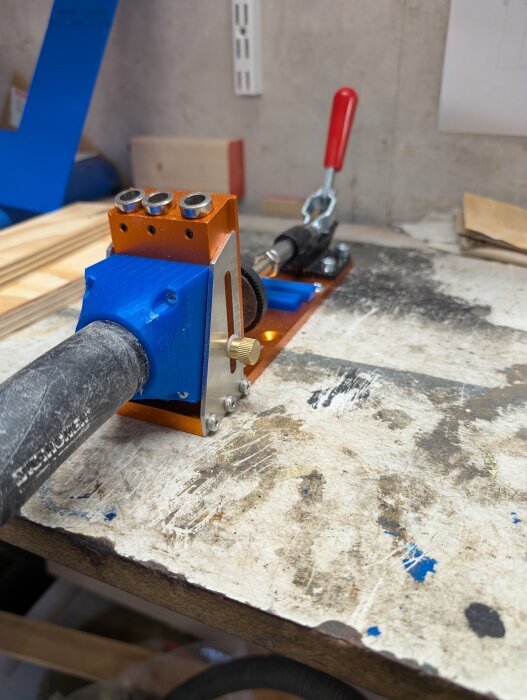 Pocket hole jig och dammsugare på arbetsbänk, med trästycken i bakgrunden och borrverktyg synligt.