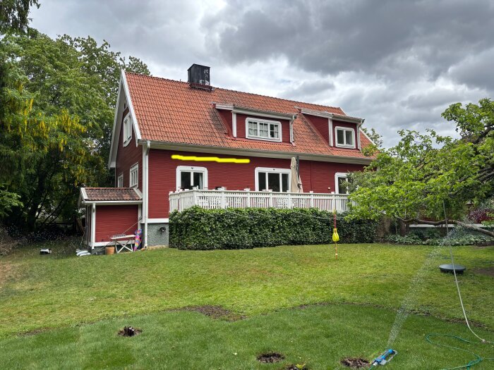 Rött hus med vita fönsterramar och staket, grönt tak och trädgård i förgrunden under en molnig himmel.