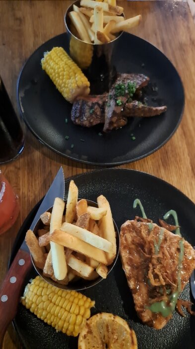 Två tallrikar med pommes frites, majs, grillat kött och kyckling, serverade på ett träbord.