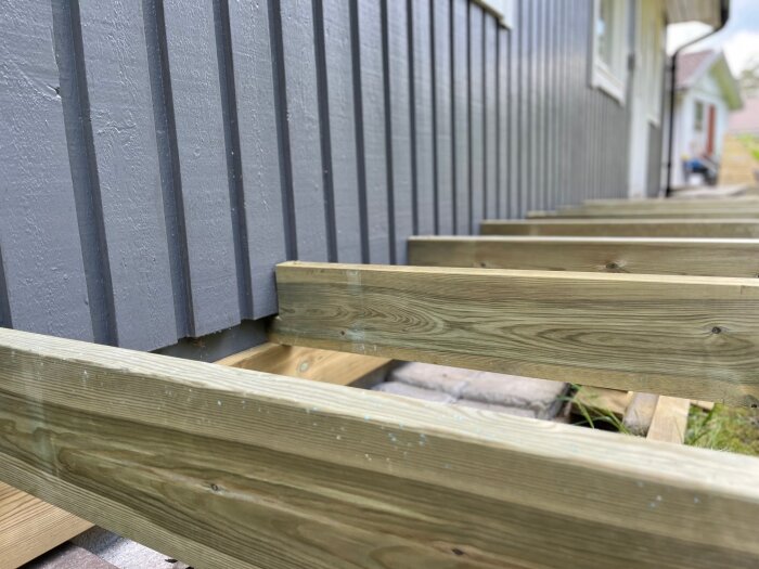 Träbalkar fästa i grunden på ett hus, visar konstruktion för altan eller veranda. Placerade parallellt längs husets grund med distans.