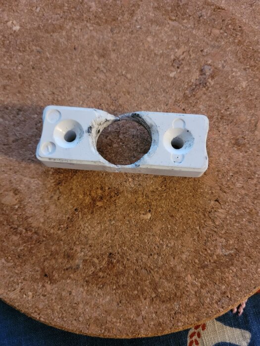 Plastdel från ett balkongdörrhandtag med två skruvhål och en stor, rund öppning i mitten, placerad på en yta av korkmaterial.