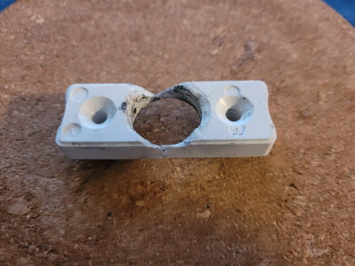 Trasig vit plastdel till dörrhandtag med två hål och en reva i mitten, placerad på en träyta.