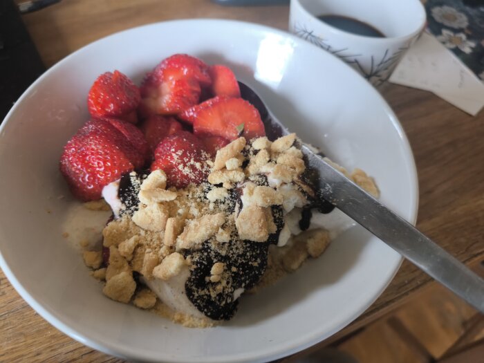 Jordgubbar, vaniljglass med chokladsås och krossade Bragokex i skål, bredvid en kopp kaffe.