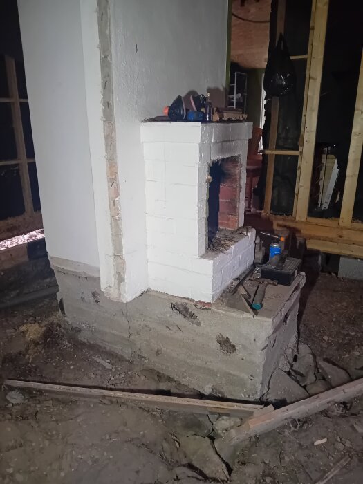 Öppen spis på murstock med spruckna tegelstenar, föremål ovanpå, och omgivande byggarbetsplats.