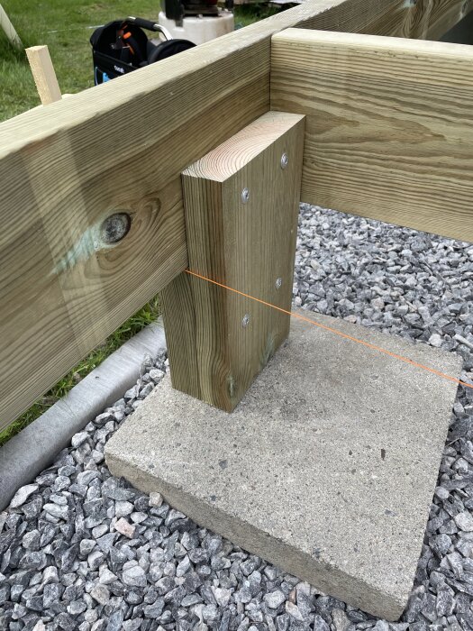 Träkonstruktion med reglar och stödben på betongplatta, temporärt stabiliserad med snöre och skruvar, omgiven av grus.