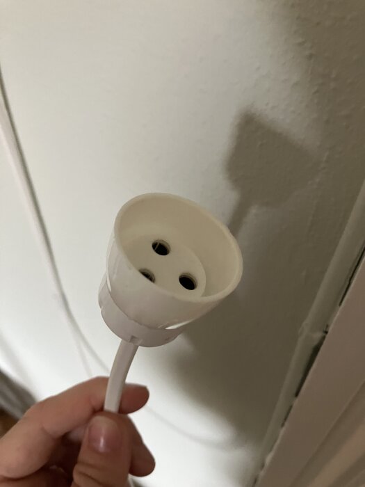 Hand håller i en vit ojordad stickkontakt till taklampa, mot en ljus vägg.