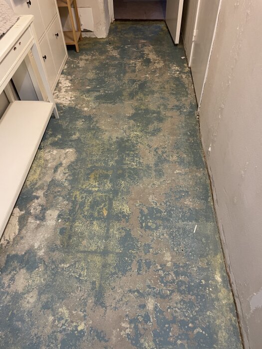 Betonggolv i källargång med fläckig yta, omgiven av vitmålade väggar och vita möbler, förberedelse inför installation av klinker.